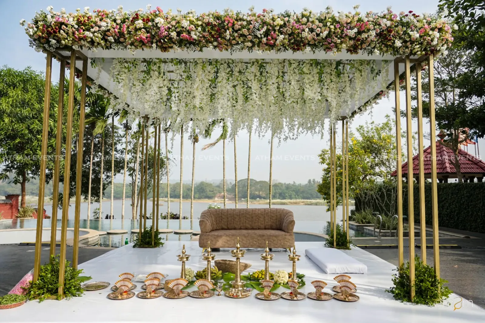 Lakefront wedding mandap with white hanging flowers and gold pillars by Atham Events